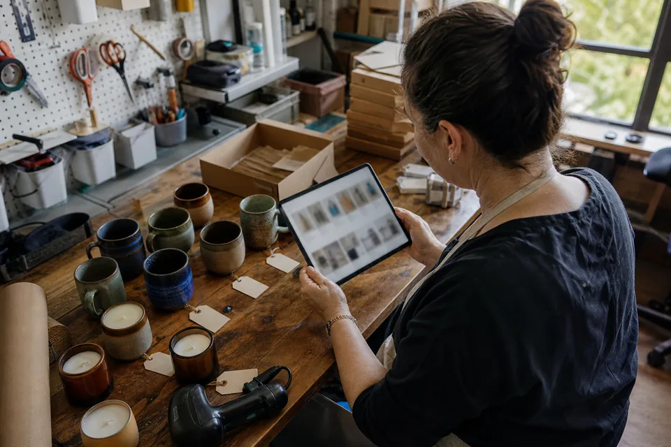 A shop owner compares product tiles on a tablet with the real products and price tags laid out on a workbench