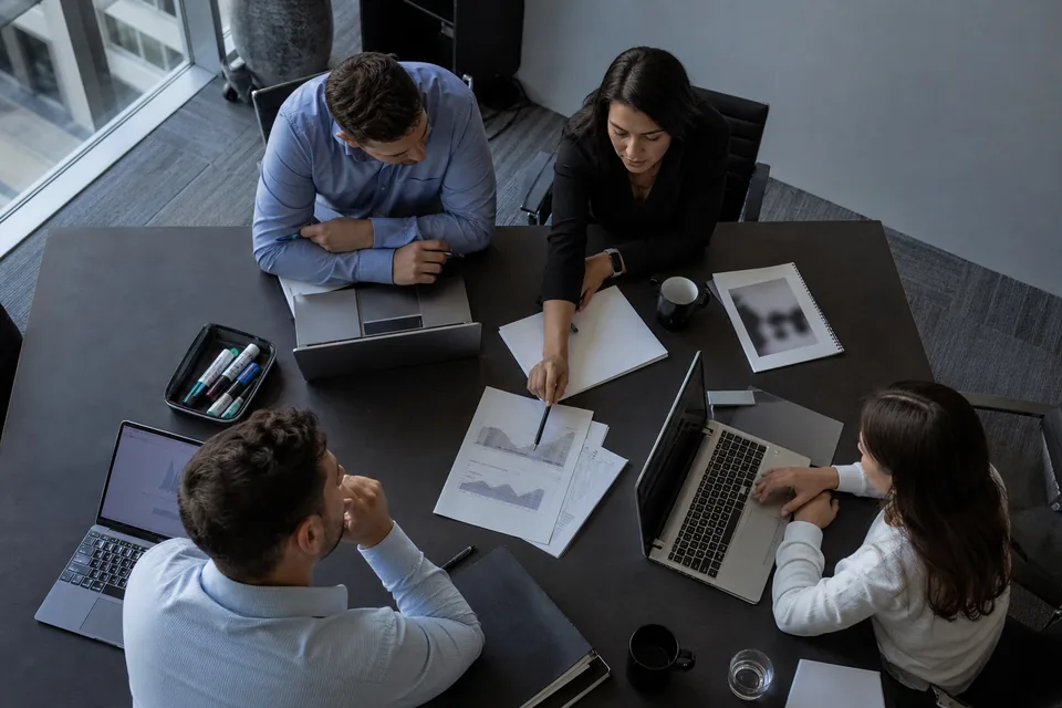Overhead view of a conference table with open laptops, printed charts and four people in a meeting