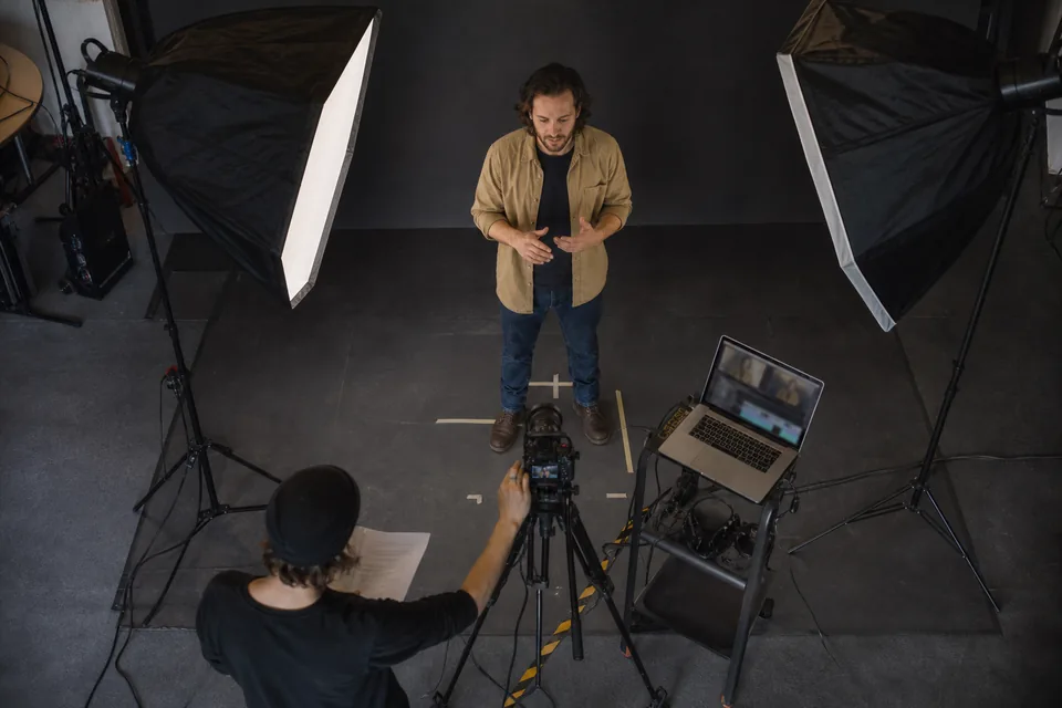 View down from a lighting grid into a video studio with camera, softboxes and a presenter on floor marks