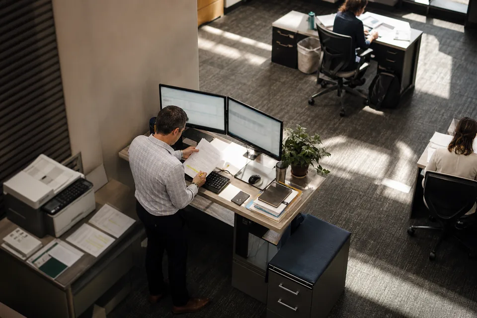 View down through a skylight into an office where a manager reviews a printed budget at a standing desk