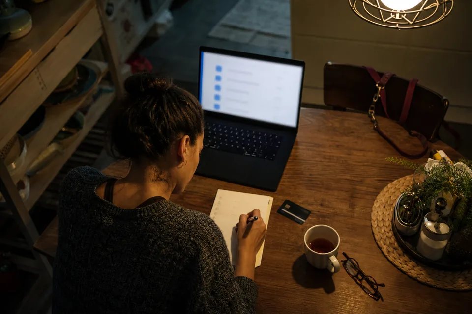 Overhead view of a kitchen table with a laptop showing an onboarding checklist, a notebook with a numbered list and a tea mug